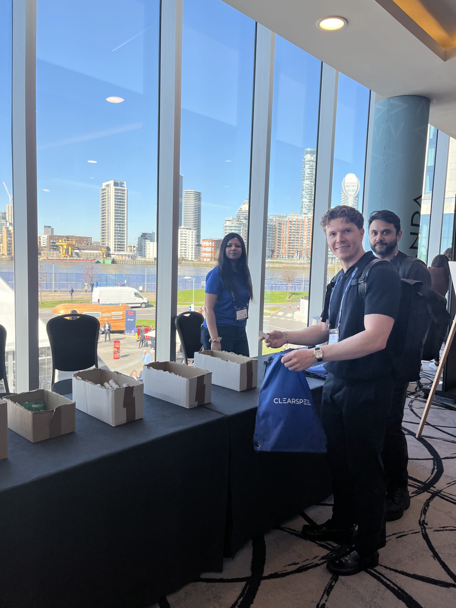 A smiling attendee holds up a blue Clearspeed hygiene kit bag at the build table, with completed kits in boxes beside him
