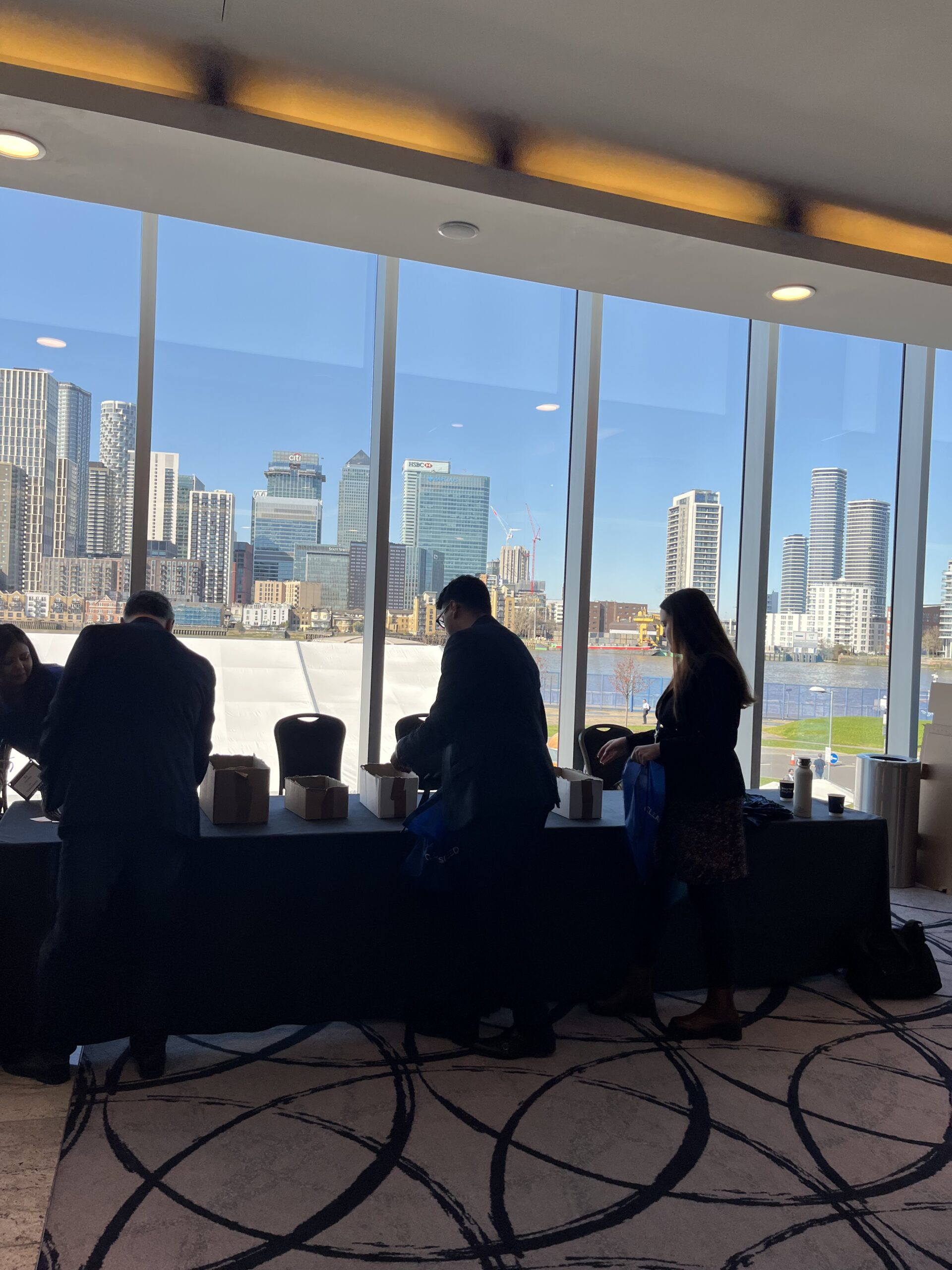 Attendees building hygiene kits at long tables in front of floor-to-ceiling windows overlooking the Thames and Canary Wharf