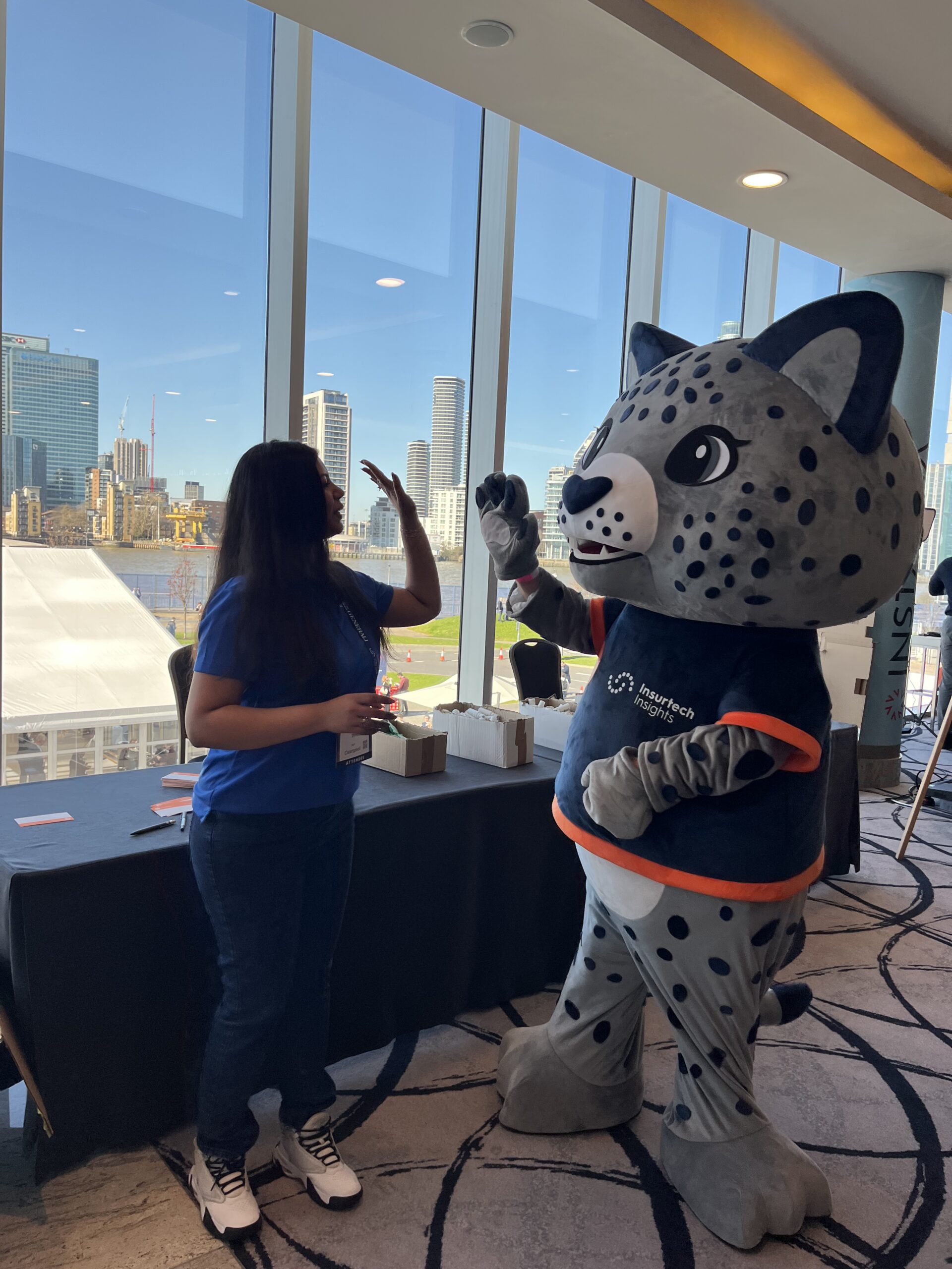 A woman at the build table and the Insurtech Insights mascot wave to each other in front of windows overlooking the Thames