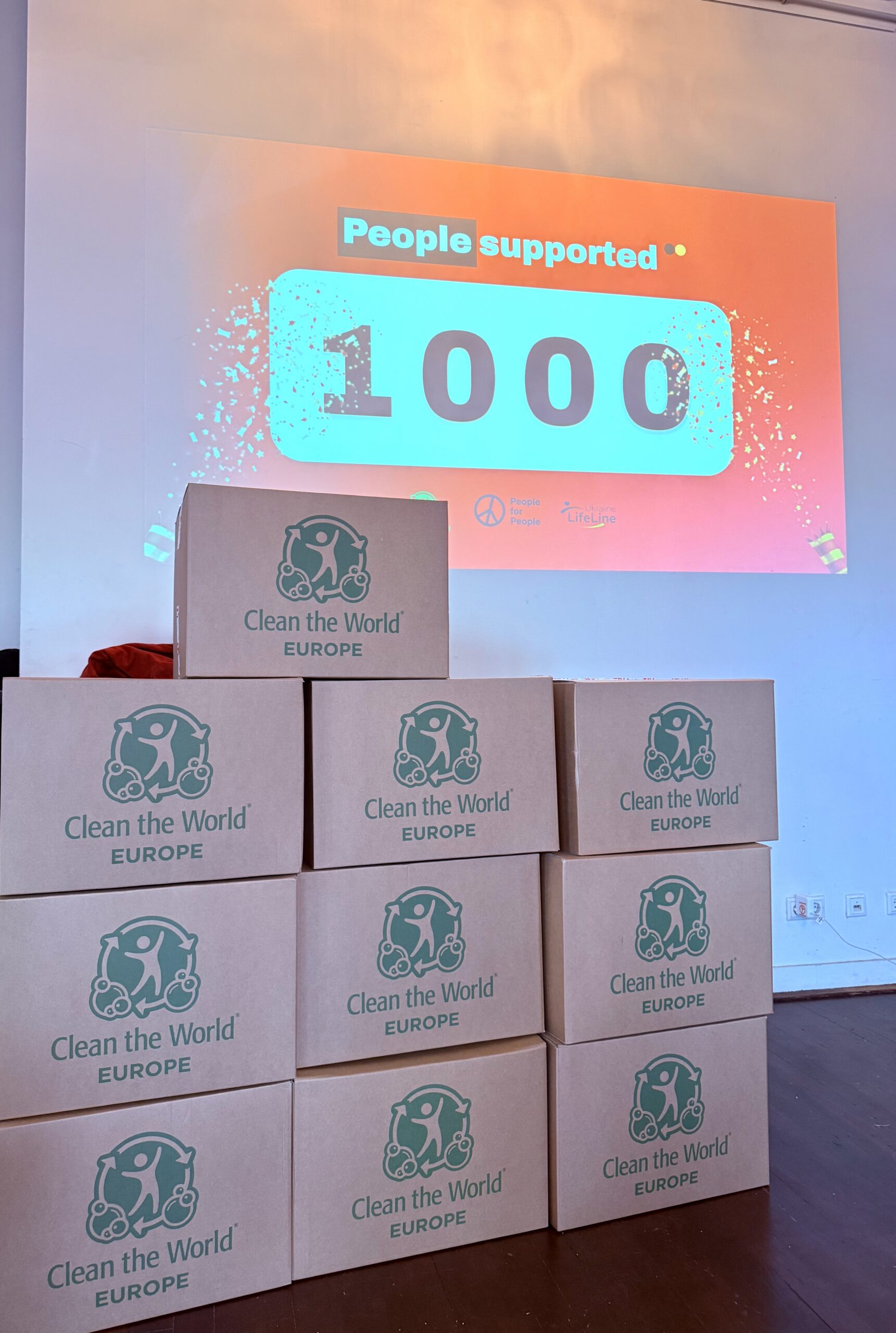 Stacked boxes of assembled hygiene kits in front of a presentation screen showing a 1,000 counter at the Young Impact Summit in Amsterdam